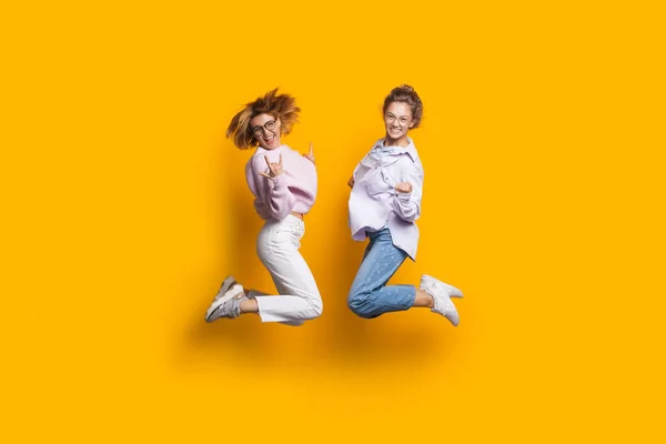 Дві блондинки-сестри стрибають на жовтій стіні студії і посміхаються на камеру жестикулюючи щастя — стокове фото