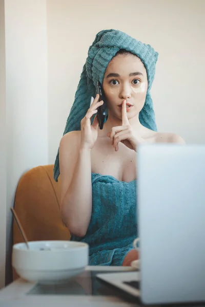 Kavkazská žena po koupeli je mluvení po telefonu a nosit hydrogel oční cestu, zatímco sedí na notebooku a jíst něco gestikulující ticho zvuk — Stock fotografie