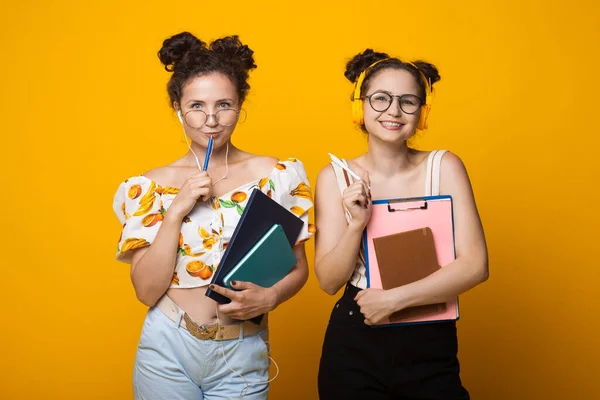 Gemelos rizados sonriendo a la cámara a través de gafas con auriculares y sosteniendo algunas carpetas en una pared amarilla — Foto de Stock