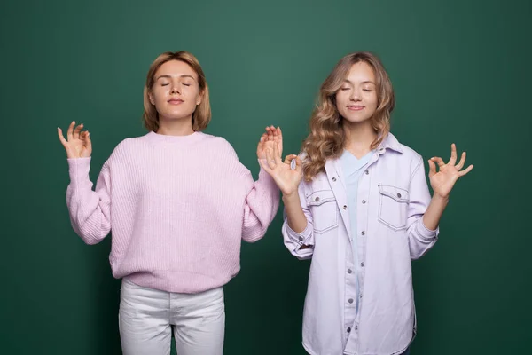 Zwei meditierende blonde Damen posieren in einer speziellen Yogaposition mit geschlossenen Augen an einer grünen Studiowand — Stockfoto