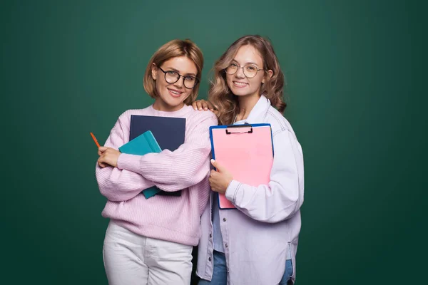 Dos estudiantes posando con algunas carpetas y sonriendo a la cámara usando anteojos en una pared verde del estudio — Foto de Stock