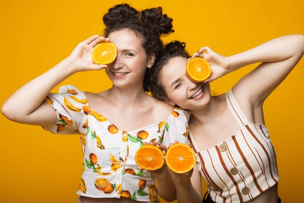 白种人双胞胎用橙子遮住他们的眼睛，对着黄色墙上的相机微笑 — 图库照片