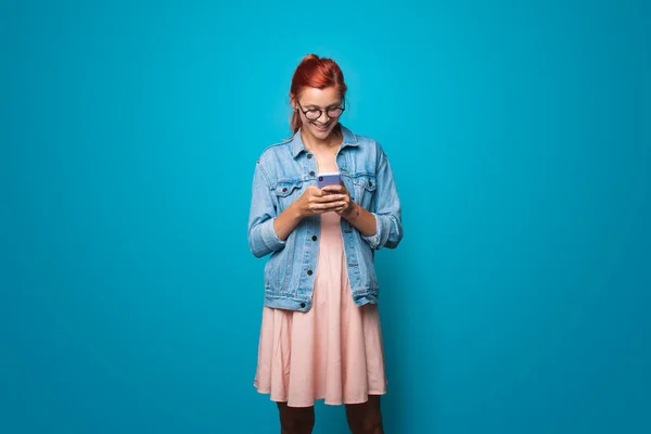 Jengibre mujer caucásica en un vestido y jeans chaqueta es sonrisa usando gafas y charlando en un teléfono en el estudio azul — Foto de Stock