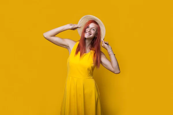 照片上的一个高加索女人，红头发，身穿连衣裙，头戴帽子，站在黄色的工作室墙上，对着相机微笑 — 图库照片