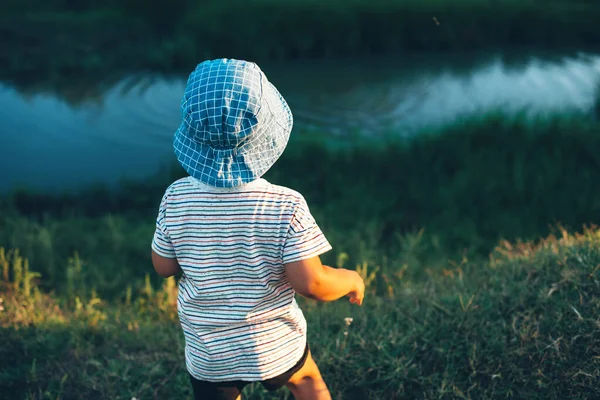 Kaukasischer Junge blickt auf einen kleinen Fluss und wirft mit einem blauen Hut Steine in ein grünes Feld — Stockfoto