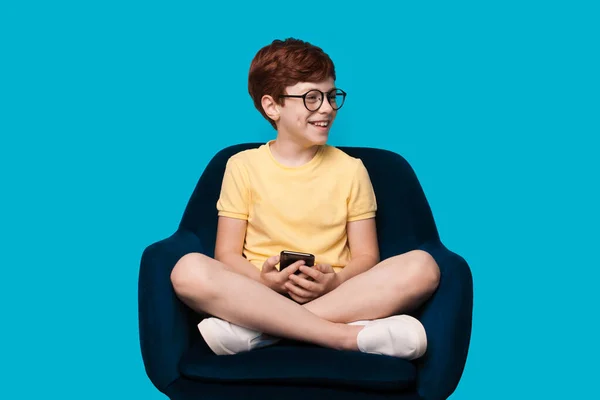 Ginger ragazzo caucasico sorridente e indossare occhiali su un muro di studio blu, mentre tenendo e utilizzando un cellulare — Foto Stock