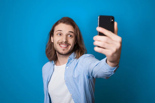 Jovem homem de cabelos longos com barba está sorrindo e fazendo uma selfie usando um telefone em uma parede de estúdio azul em roupas casuais — Fotografia de Stock