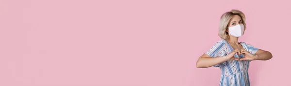 Wanita pirang lucu mengenakan masker medis dan pakaian adalah tanda hati gestur di dinding merah muda dengan ruang kosong — Stok Foto