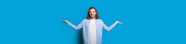 青い壁の上でフリースペースでポーズヤシの上に2つの事を比較し、バランスをとることによって何かを宣伝しているひげの長い髪の男 — ストック写真