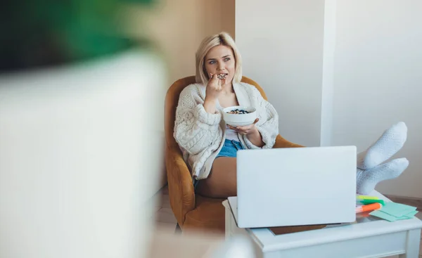 Rubia mujer caucásica con un ordenador portátil está comiendo cereales mientras tiene clases en línea — Foto de Stock