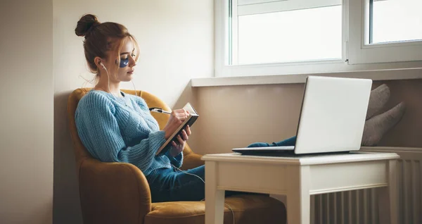 Kızıl saçlı ve çilli beyaz bir kadın dizüstü bilgisayar kullanırken ve hidrojel göz bandı takarken bir kitaba bir şeyler yazıyor. — Stok fotoğraf
