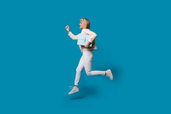 Laufende blonde Frau hält eine Geschenkbox an einer blauen Studiowand und lächelt in lässiger Kleidung — Stockfoto