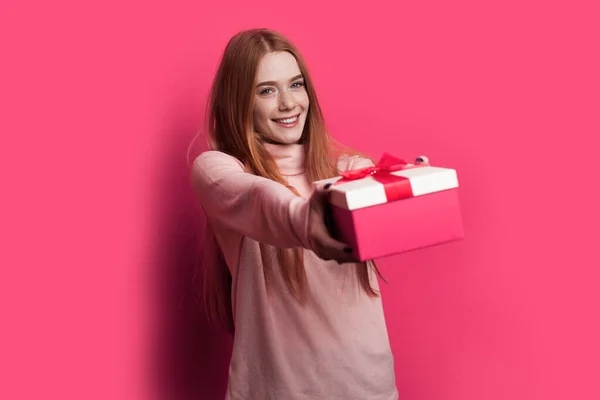 Mujer pelirroja con pecas y cabello rojo sonríe y da a la cámara una caja roja con regalo posando en una pared roja — Foto de Stock