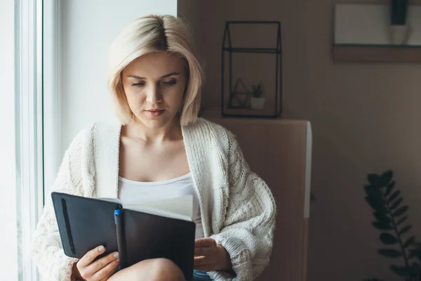 Rubia mujer caucásica vistiendo un suéter de punto está leyendo un libro sentado cerca de la ventana en casa — Foto de Stock