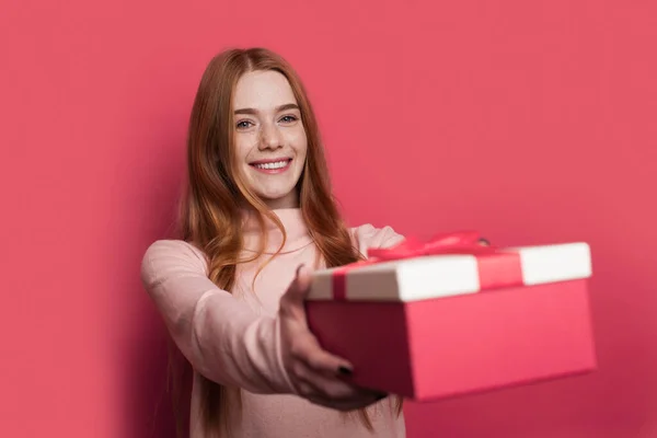 Joven mujer encantadora con el pelo rojo está dando a la cámara un regalo sonriendo en una pared de estudio rojo — Foto de Stock