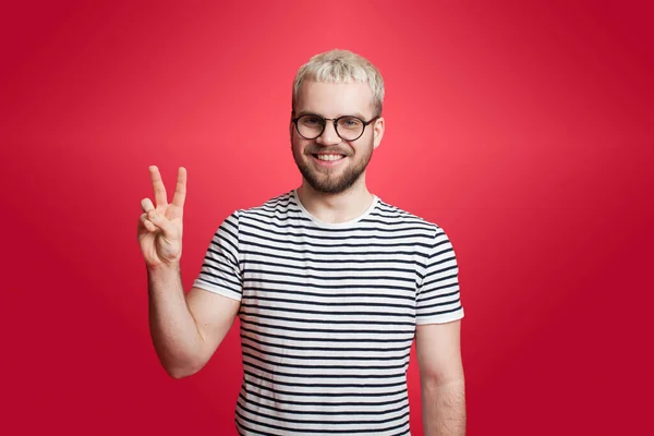 Homem caucasiano com cabelo loiro e barba está gesticulando o sinal de paz em uma parede vermelha sorrindo para a câmera — Fotografia de Stock