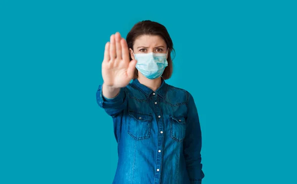 Serieuze blanke vrouw met medisch masker op gezicht is gebaren een stopbord met palm poseren op een blauwe studio muur — Stockfoto