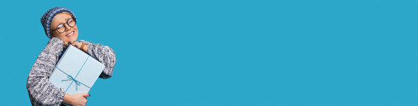 Блондинка в шляпе и очках крепко обнимает подарок, рекламируя что-то на голубой стене с бесплатным пространством