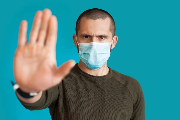 Close-up foto van een blanke man in trui met een medisch masker op een blauwe studiomuur gebaren stop en afwijzing bord met palm — Stockfoto