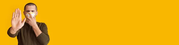 Homme caucasien aux cheveux courts avec masque médical sur le visage est gestuelle stop signe avec la paume posant sur un mur jaune avec espace libre — Photo
