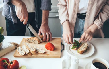 Beyaz bir çift mutfakta ekmek ve sebze doğrarken birlikte sandviç hazırlıyor.