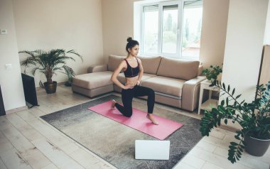 Beyaz kadın spor giyimli, pembe halıda dizüstü bilgisayar kullanırken evde spor egzersizleri yapıyor.