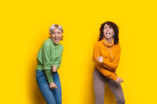 Verrückte Frauen mit Kopfhörern tanzen in lässiger Kleidung auf einer gelben Studiowand und lächeln in die Kamera — Stockfoto