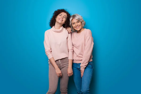 Schöne blonde und brünette Schwestern posieren auf einer blauen Wand im Studio in denselben Pullovern und Jeans und lächeln fröhlich in die Kamera — Stockfoto