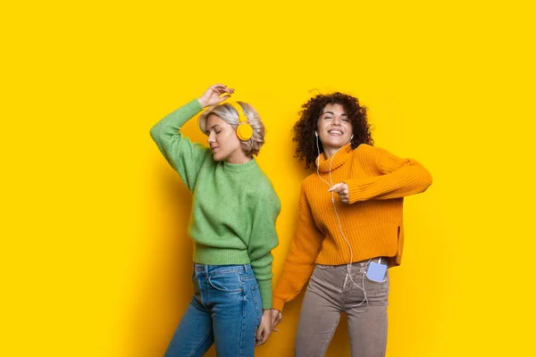 Donne affascinanti con i capelli ricci ballano ascoltando musica in cuffia su una parete gialla dello studio indossando maglioni caldi e sorridendo. — Foto Stock
