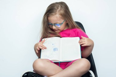 Beyaz bir stüdyo duvarındaki koltukta oturan sarı saçlı ve gözlüklü küçük beyaz kız kameraya el kol hareketleriyle kitabını çiziyor.