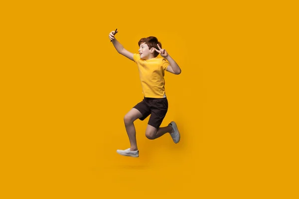 Ginger kaukaski chłopiec skacze na żółtej ścianie studio i robi selfie za pomocą telefonu gestykulując znak kawałek i uśmiech — Zdjęcie stockowe