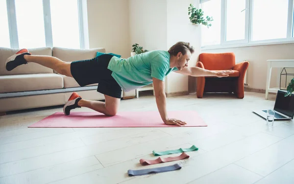 Hombre caucásico con cabello rubio usando ropa deportiva está planchando y estirándose en una alfombra de yoga rosa en casa usando una computadora portátil — Foto de Stock