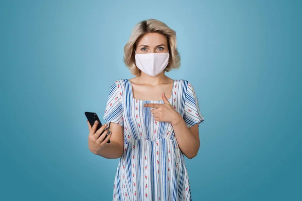 Wanita pirang kaukasia dengan topeng medis di wajah memegang telepon dan menunjuk pada dinding studio biru — Stok Foto