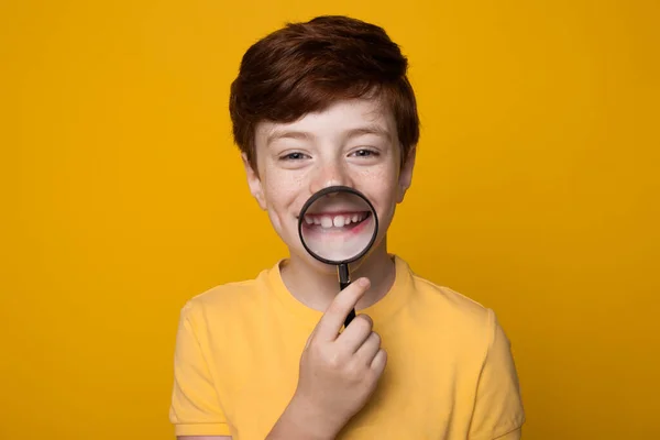 Gengibre caucasiano menino está segurando uma lupa em sua boca e sorrir toothy na câmera posando em uma parede de estúdio amarelo — Fotografia de Stock