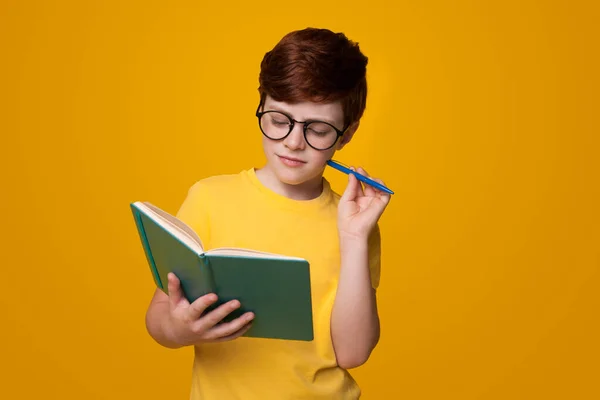 Skoncentrowany biały chłopiec czyta książkę na żółtej ścianie studia trzymając długopis i nosząc okulary. — Zdjęcie stockowe