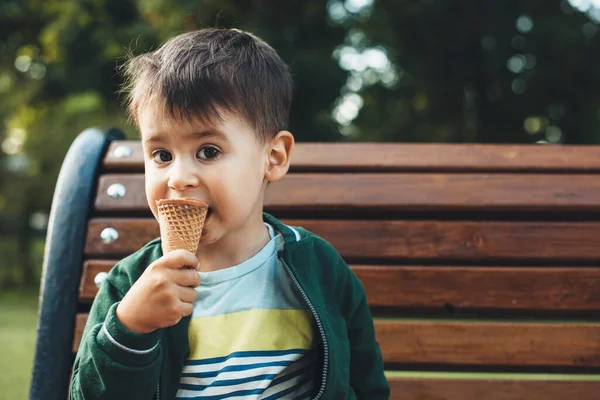 Adorável menino caucasiano pequeno comer um sorvete enquanto sentado em um banco no parque — Fotografia de Stock