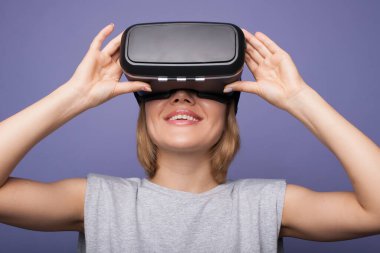 Sarışın kadın bir vr kulaklık takıyor ve menekşe bir stüdyo duvarında bir şey izlerken gülümsüyor.