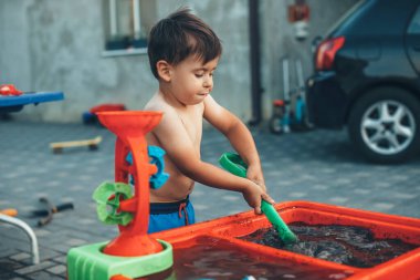 Küçük beyaz çocuk bahçede plastik su oyuncaklarıyla oynuyor. Hem de bir yaz günü çıplak karnıyla.