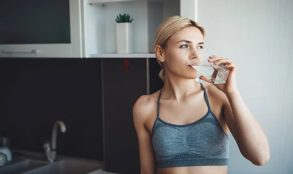 Lovely caucasian woman with blonde hair wearing sportswear is drinking a glass of water during the digital fitness lessons at home — Stock Photo, Image