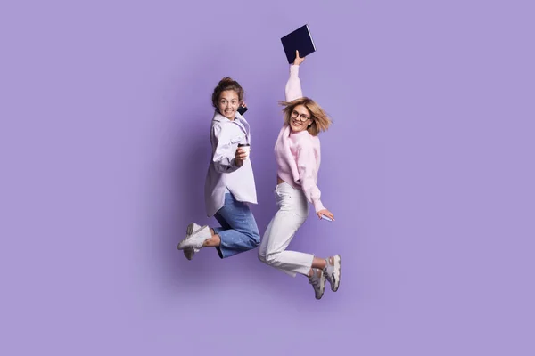 Dos estudiantes caucásicos en ropa casual están saltando en una pared de estudio violeta mientras sostienen un libro y sonríen a la cámara — Foto de Stock