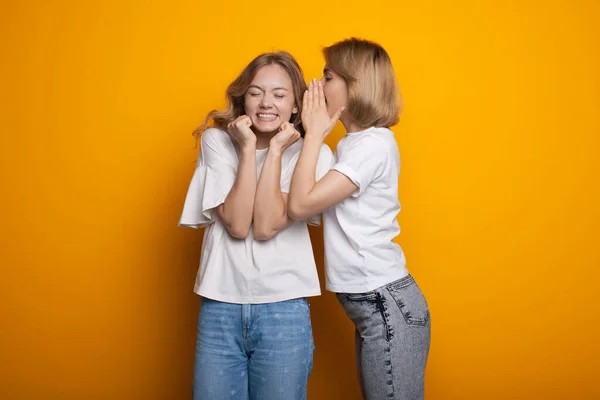 Blonde Kaukasierin flüstert ihrem Freund etwas zu, posiert in lässiger Kleidung auf einer gelben Studiowand — Stockfoto