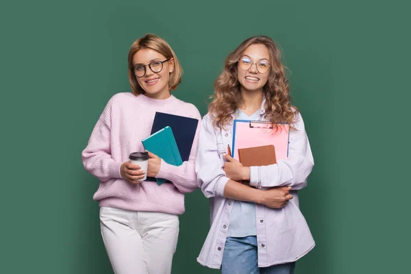 Charmante blonde Studenten lächeln an einer grünen Wand, während sie ein paar Bücher umarmen und einen Kaffee trinken — Stockfoto