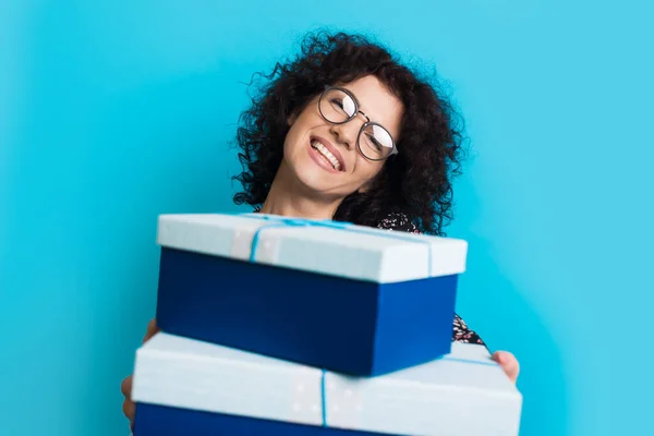 Vit kvinna med lockigt hår och glasögon ger en present vid kameran ler på en blå ateljé vägg — Stockfoto