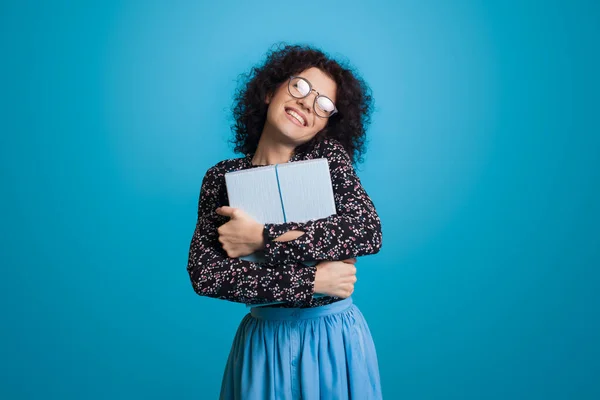 Słodka biała kobieta z kręconymi włosami i okularami obejmuje pudełko z prezentami uśmiechnięte na niebieskiej ścianie studia w sukience — Zdjęcie stockowe
