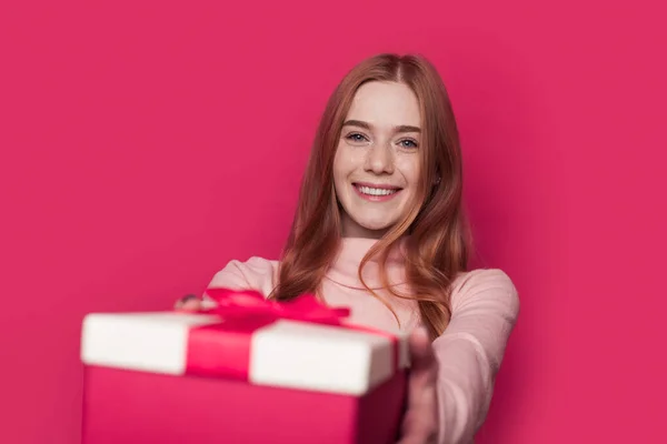 Encantadora mujer de jengibre con pecas está dando un regalo a la cámara sonriendo en una pared rosa en el estudio — Foto de Stock