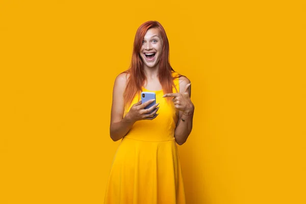 Foto monocromática de una mujer caucásica con un vestido y el pelo rojo apuntando a su teléfono en una pared de estudio amarillo — Foto de Stock