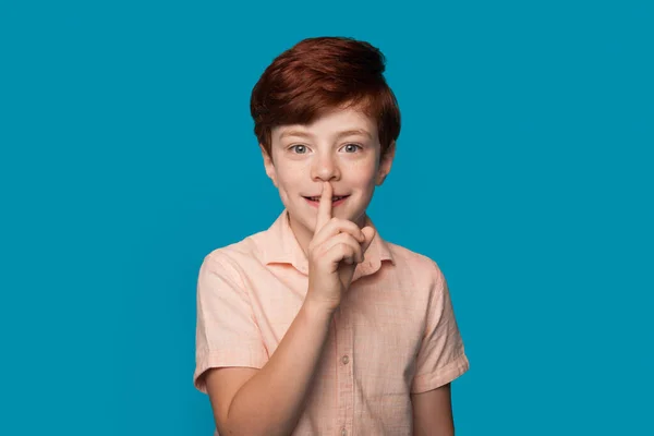 Gengibre caucasiano menino está gesticulando o silêncio som sorrindo em uma parede de estúdio azul em roupas casuais — Fotografia de Stock
