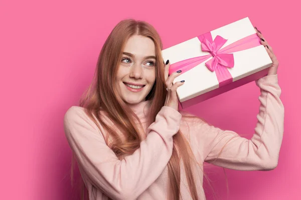 Preciosa mujer de jengibre con pecas y el pelo rojo está sacudiendo un regalo y sonrisa en una pared de estudio rosa — Foto de Stock