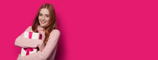 Cute red haired woman is embracing a present and smiling at camera on a pink wall with free space — Stock Photo, Image