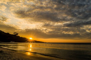 Bulutlu gökyüzünün altında denizde ve sahilde gün batımı.